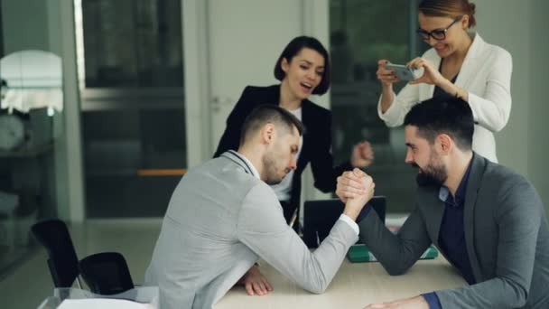 Handsome guys in trendy suits are competing in arm wrestling sitting at desk in office while girls are recording them using smartphone camera then doing high-five. - Filmagem, Vídeo