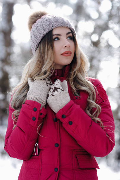 Beautiful woman standing among snowy trees in winter forest and enjoying first snow. Wearing knitted hat whith ball and red down jacket.  Winter holiday concept - Image - Foto, Imagem