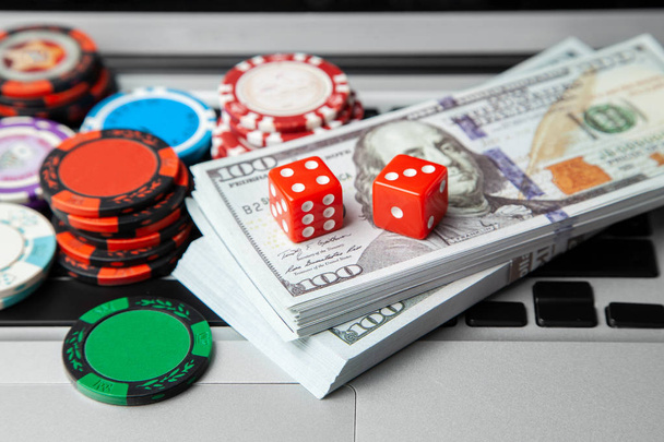オンラインカジノのノート パソコン。ノート パソコンのキーボードと緑のゲーム テーブルのサイコロとお金の現金ドルのチップ。ゲーム中毒ギャンブル - 写真・画像