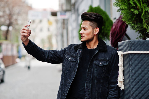 Bonito e elegante homem indiano em jaqueta de jeans preto posou ao ar livre e fazendo selfie no telefone
. - Foto, Imagem