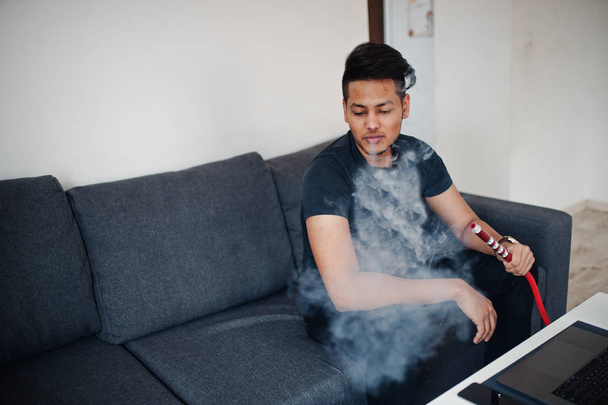 Handsome and fashionable indian man in black sitting at room, smoking hookah - 写真・画像