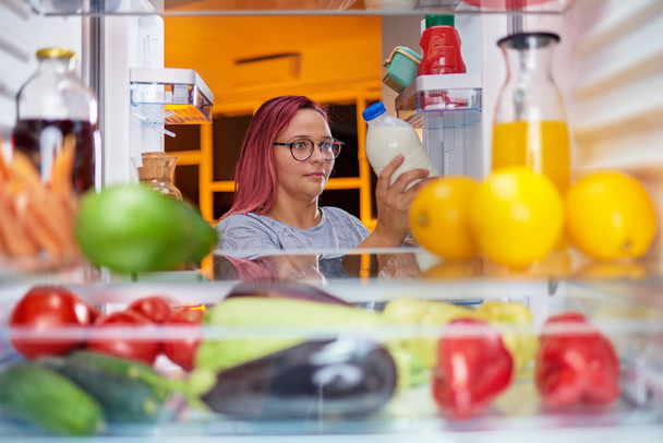 Thick woman taking milk from fridge full of groceries. Unhealthy eating, eating disorder. Picture taken from inside of fridge. - Valokuva, kuva