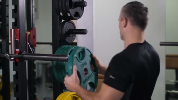 Handsome, strong man puts a disk on the barbell, turns to the camera and smiles 4K Slow Mo - Footage, Video
