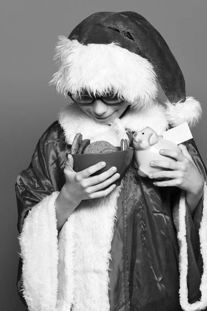 jeune garçon santa claus mignon avec des lunettes en pull rouge et chapeau de Noël nouvel an tenant tirelire rose et biscuits de la hanche au chocolat dans un bol sur fond de studio bleu
 - Photo, image