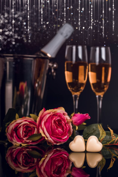 Μπουκάλι σαμπάνιας σε κουβά με δύο ποτήρια κρασί, μπουκέτο με τριαντάφυλλα και καρδιά σχήμα σοκολάτες με φώτα σε μαύρο φόντο. Αγάπη, ημέρα του Αγίου Βαλεντίνου έννοια - Φωτογραφία, εικόνα