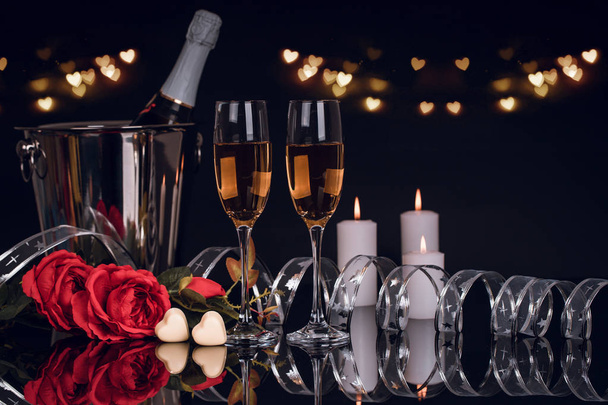 Butelka szampana w wiaderku z dwa kieliszki do wina, bukiet róż, czekoladki w kształcie serca i świece ze światłami na czarnym tle. Miłość, Walentynki koncepcja - Zdjęcie, obraz