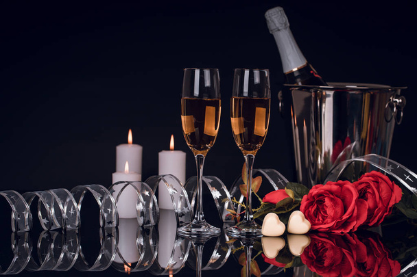 Champagne bottle in bucket with two wine glasses,  bouquet of  roses,  heart shaped chocolates and candles on black background. Love, Valentines day concept - Photo, Image