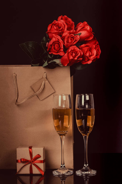 Champagne bottle in a paper bag, two wine glasses and  red roses on black background. Love, Valentines day concept - Photo, Image