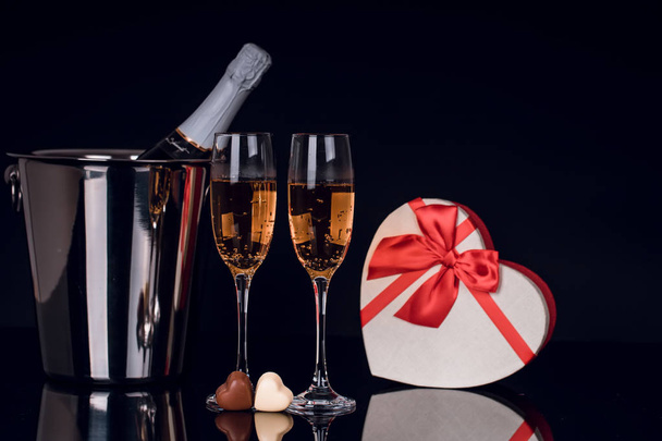 Champagne bottle in bucket, two wine glasses, two heart-shaped chocolates and gift box on black background. Love, Valentines day concept - Photo, Image