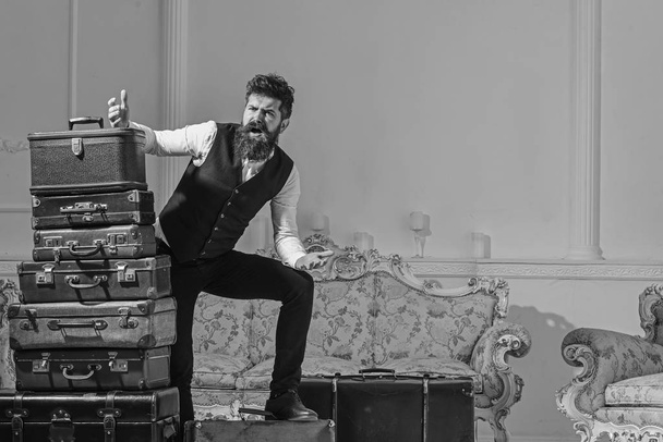 Man with beard and mustache packed luggage, white interior background. Move out, transportation concept. Macho elegant on desperate face, exhausted at end of packing, near pile of vintage suitcases - Φωτογραφία, εικόνα