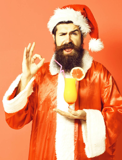 handsome bearded santa claus man with long beard on happy face holding glass of nonalcoholic cocktail in christmas or xmas sweater and new year hat showing ok on red studio background - Valokuva, kuva