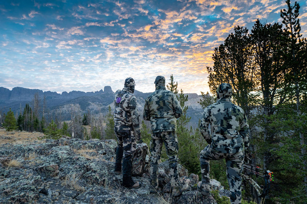 Tres amigos cazadores adultos, irreconocibles, se paran en una cresta de montaña buscando alces para cazar durante la temporada de tiro con arco. Usar camuflaje
 - Foto, imagen