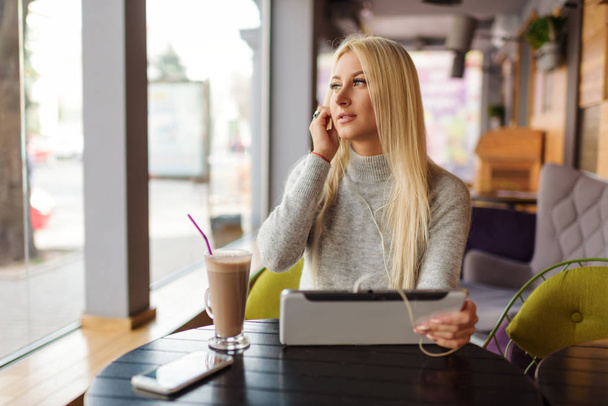 Young blond Woman using Tablet and Drinking Coffee. Relaxing in Cozy cafe, Sit on the Chair and listening with pleasure in headphones an audio book or music - Photo, image