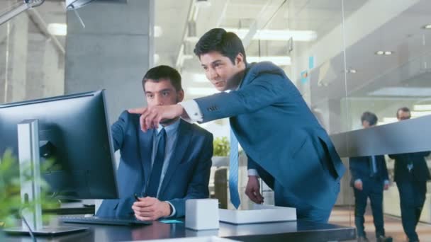 В офисе бизнесмена, сидящего за своим столом, беседующего со своим коллегой, они указывают на экран компьютера. Профессиональные бизнесмены в стильном офисе
. - Кадры, видео
