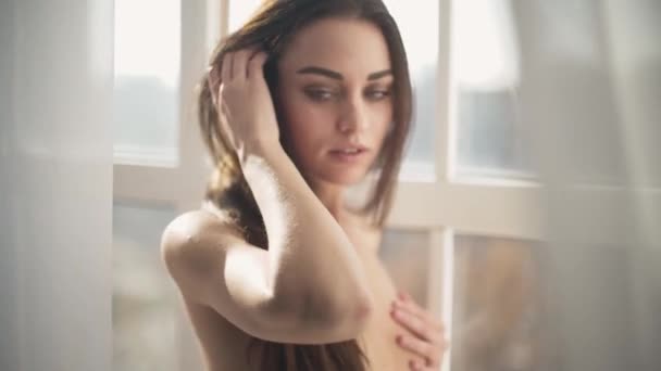 Portret van een mooie sensuele naakt meisje met donkere haren, mooie ogen en tedere lippen staande aan de voorkant van een brede helder venster. - Video