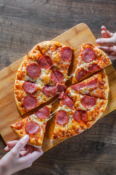 Zwei Frauen nehmen eine Scheibe Paprika-Pizza mit Mozzarella-Käse, Salami, Tomaten, Pfeffer. italienische Pizza auf Holztischhintergrund - Foto, Bild