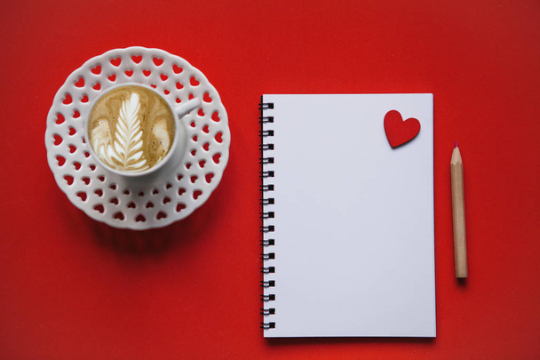 Tuoretta cappuccino-kahvia punaisella pohjalla. Lähellä tyhjää päiväkirjaa sydämellä kirjoittaa
. - Valokuva, kuva