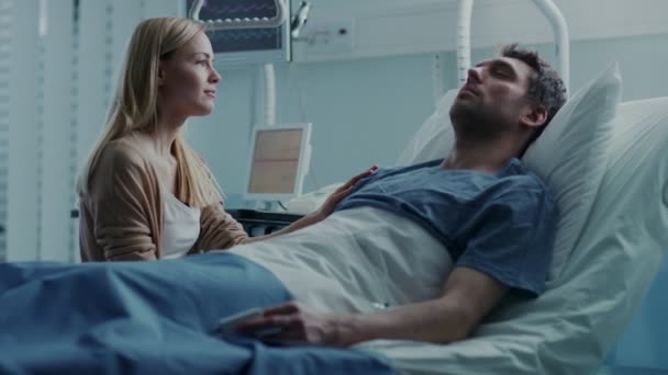 In the Hospital Sick Man Lying on the Bed, His Visiting Wife Hopefully Sits Beside Him and Prays for His Rapid Recovery. Tragic, Somber and Melancholy Scene. - Felvétel, videó