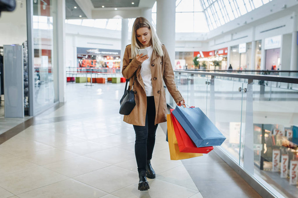 Girl looks at something in her phone walking around the big mall with many stores with shopping bags on her shoulder. Consumerism and lifestyle concept - Image - 写真・画像