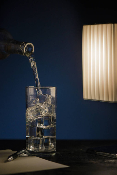 A glass of sparkling water with ice on a dark table with a pen and a sheet of paper and a desk lamp. - Foto, Imagen