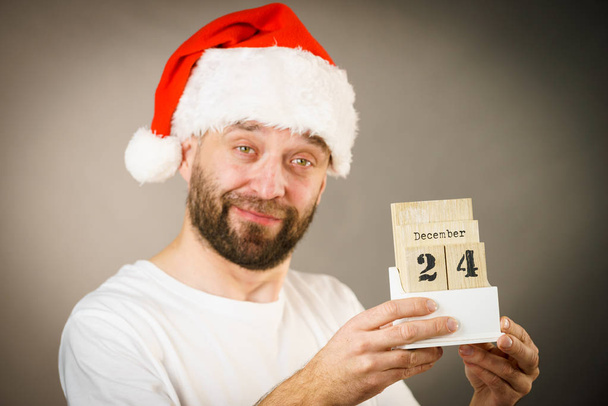 Man wearing santa claus christmas hat holding calendar with 24 december date, on grey - Photo, image