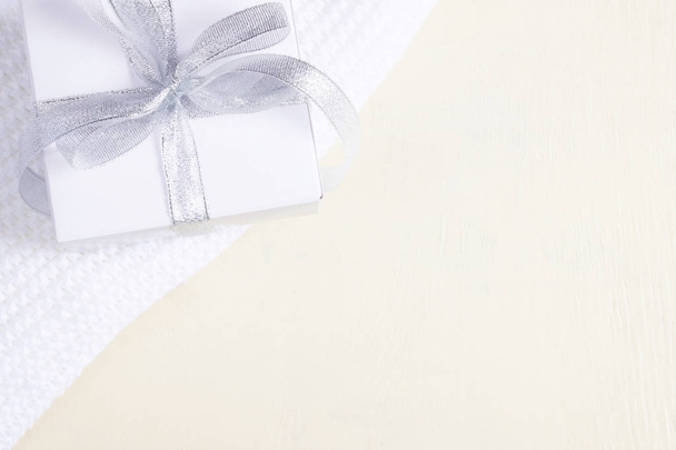 Composition of white beautiful gift box with silver ribbon on warm hand-knitted blanket on wooden painted background. Flat lay style with copy space. - Photo, image