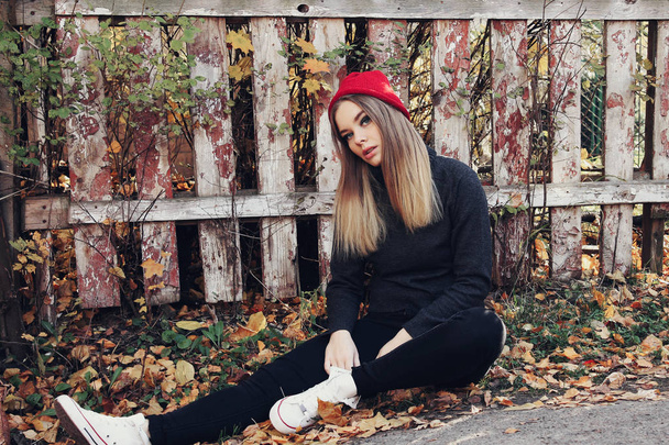 Cool chica de moda en un sombrero rojo se apoya contra una pared de la cerca. Estilo Hipster. Moda juvenil
. - Foto, Imagen