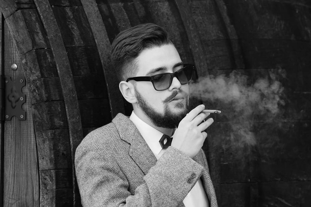 Jonge brutale man met baard in zonnebril roken van een sigaret in stad straat. Stijlen mode mannelijk model in jas - Foto, afbeelding