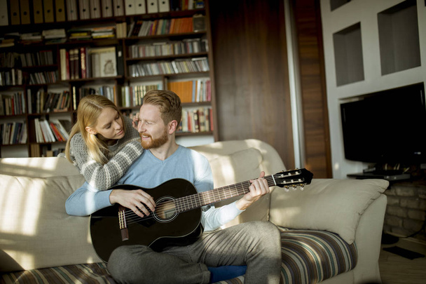 Homem tocando guitarra acústica no sofá para sua jovem mulher bonita
 - Foto, Imagem