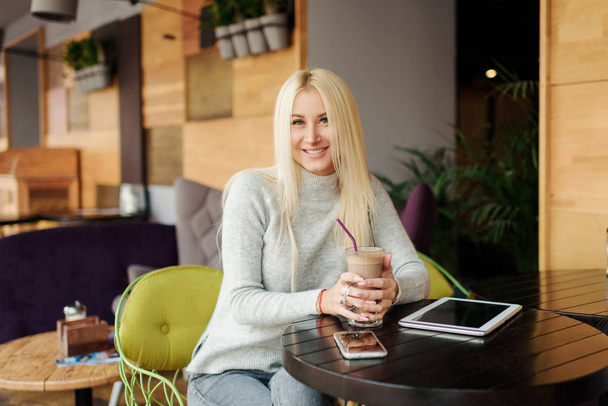 Young blond Woman using Tablet and Drinking Coffee. Relaxing in Cozy cafe, Sit on the Chair and listening with pleasure in headphones an audio book or music - Fotografie, Obrázek