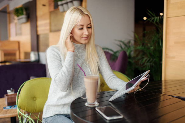 Young blond Woman using Tablet and Drinking Coffee. Relaxing in Cozy cafe, Sit on the Chair and listening with pleasure in headphones an audio book or music - Photo, Image