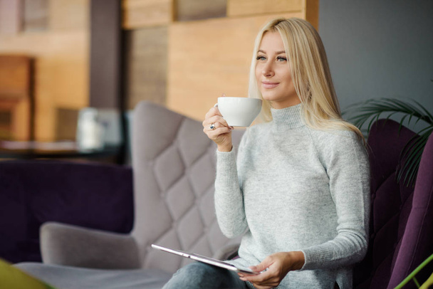 Reading Concept. Young blond Woman using Tablet and Drinking Coffee. Relaxing in Cozy cafe, Sit on the Chair - Фото, изображение
