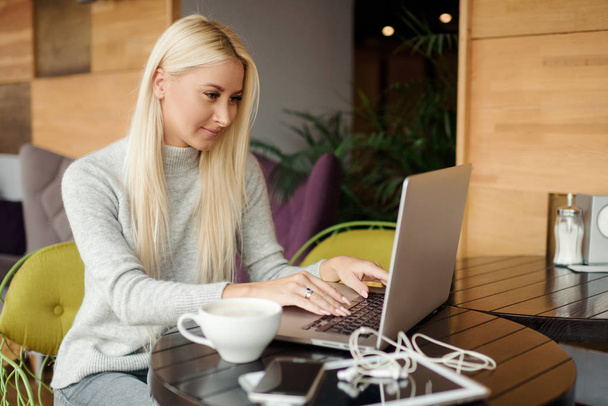 Concentrated at work. Confident young woman in smart casual wear working on laptop while sitting near window in creative office or cafe. Holding a cup of coffee - Foto, Imagem