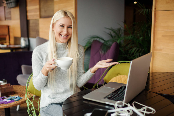 Концентрований на роботі. Впевнена молода жінка в розумному повсякденному одязі працює на ноутбуці, сидячи біля вікна в творчому офісі або кафе. Тримаємо чашку кави
 - Фото, зображення