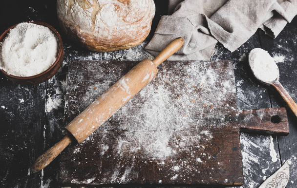 baked bread, white wheat flour, wooden rolling pin and old cutting board on a black table, top view - Fotografie, Obrázek