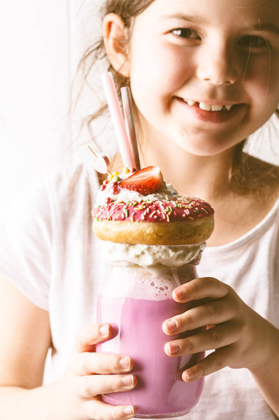 Little smiling girl drinks pink strawberry freak shake cocktail with donut and sweets, white background. Unhealthy desserts concept. - Fotó, kép