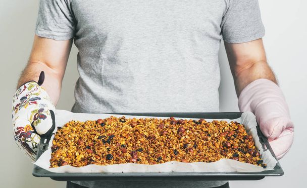 Close view of man holding holding baking tray with homemade granola from oatmeal, almonds, cashews, peanuts, hazelnuts, sunflower seeds on light background  - Foto, Bild