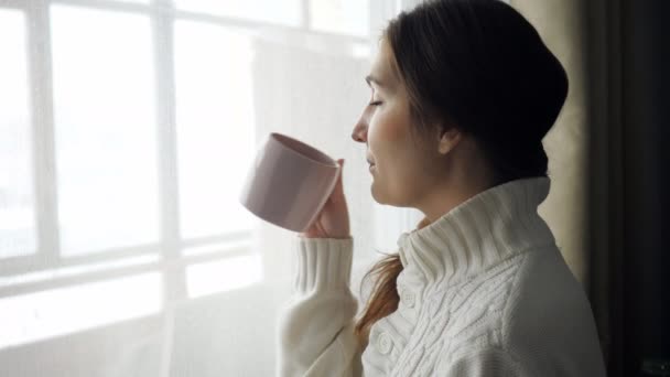 junge schöne brünette Frau im Pullover trinkt Tasse Kaffee am Fenster - Filmmaterial, Video