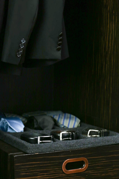 in a closet clothes, there are many ties, belts put on a tray and suits hanging on a rack - Photo, Image