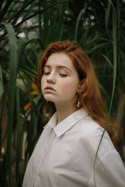 sensual red-haired woman in white shirt posing between green plants  - Foto, Imagen