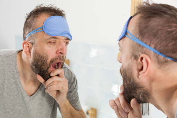 Funny adult man wearing eyemask on forhead having troubles with waking up. Standing in front of mirror looking at his face, yawning, being tired and sleepy. - Photo, Image