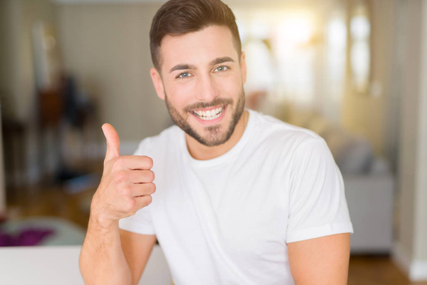 Young handsome man wearing casual white t-shirt at home doing happy thumbs up gesture with hand. Approving expression looking at the camera with showing success. - Photo, image