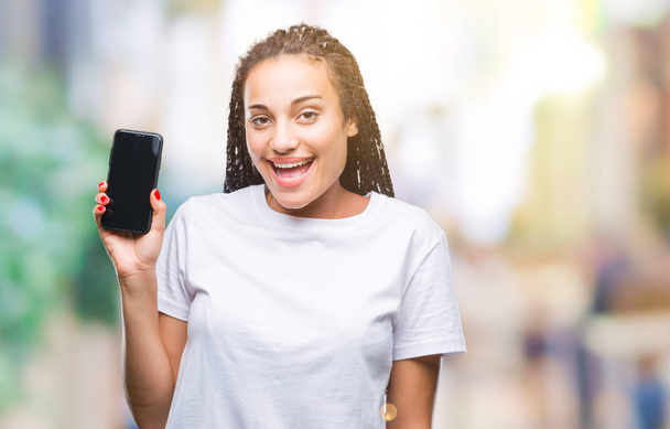 Jonge gevlochten hair Afrikaans Amerikaans meisje tonen scherm van de smartphone over geïsoleerde achtergrond met een blij gezicht permanent en lachend met een vertrouwen glimlach weergeven tanden - Foto, afbeelding
