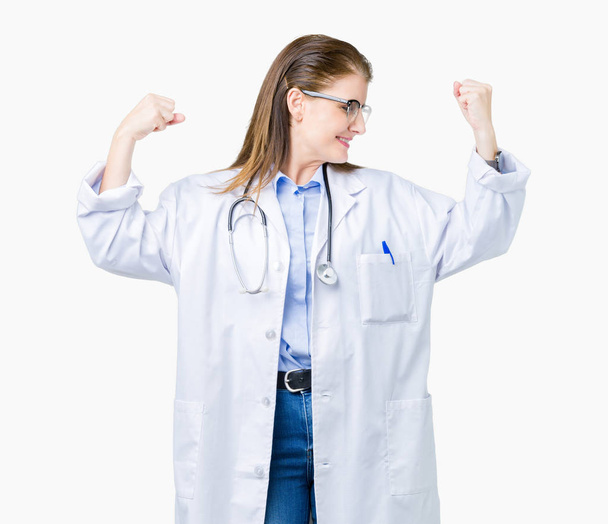 Μέσης ηλικίας γυναίκα γιατρός φορώντας ιατρική παλτό πέρα από το απομονωμένο υπόβαθρο δείχνει όπλα μύες χαμογελώντας υπερήφανος. Γυμναστήριο έννοια. - Φωτογραφία, εικόνα