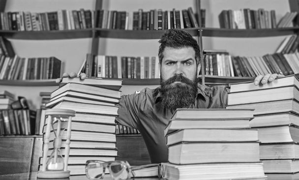 Teacher or student with beard sit at table with hourglass and glasses, defocused. Man on strict face sit between piles of books, while studying in library, bookshelves on background. Bookworm concept - Foto, immagini