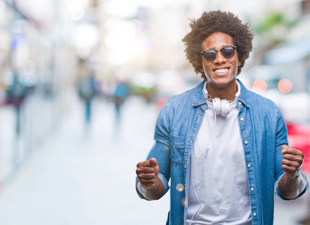 Hombre afroamericano con auriculares escuchando música sobre un fondo aislado muy feliz y emocionado haciendo un gesto ganador con los brazos levantados, sonriendo y gritando por el éxito. Concepto de celebración
. - Foto, imagen