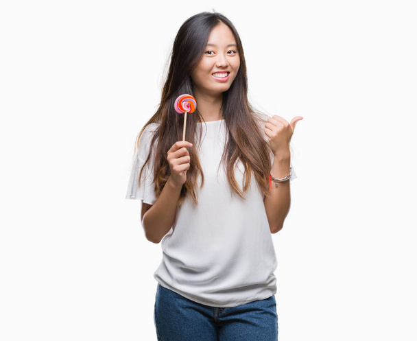 Молода азіатка їсть цукерки льодяника над ізольованим тлом, вказуючи і показуючи з великим пальцем в сторону з щасливим обличчям посміхаючись
 - Фото, зображення