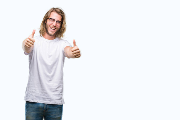 Mladý pohledný muž s dlouhými vlasy nosíš brýle izolované pozadí schvalování dělá pozitivní gesto rukou, palec se usmívá a přát úspěch. Při pohledu na fotoaparát, vítěz gesto. - Fotografie, Obrázek