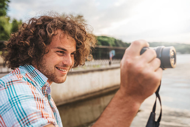 Side view portrait of young smiling photographer with curly hair photographing outdoors. Caucasian man taking a photos with digital camera outside, nature photography. People, travel and lifestyle concept. - Фото, изображение