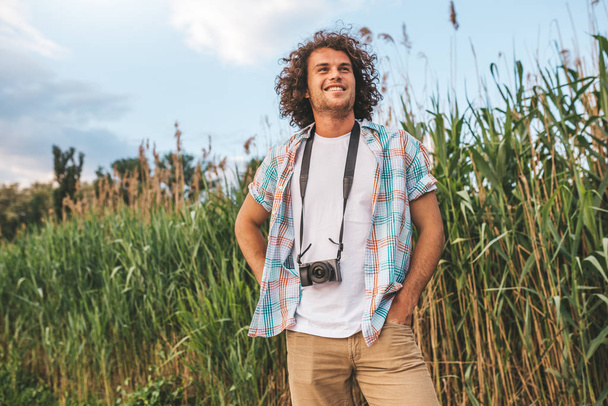 Cheerful smiling young handsome male wearing in casual with curly hair, with digital camera to take some photos of nature in vacation trip, looking a side. Travel, people and lifestyle concept. - Zdjęcie, obraz